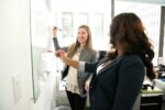 Zwei Mitarbeiterinnen im Büro stehen an der Planungstafel und besprechen Ihre Projekte